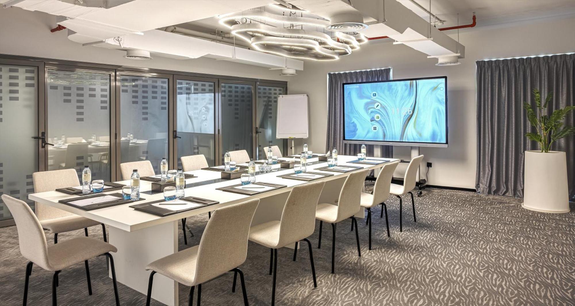 Millennium Atria Business Bay Aparthotel Dubai Luaran gambar The photo shows a modern conference room set up for a meeting. There is a long table with several chairs arranged around it, each with a notepad and pen. A large screen displays a graphic or presentation, and there is a whiteboard in the corner. The 