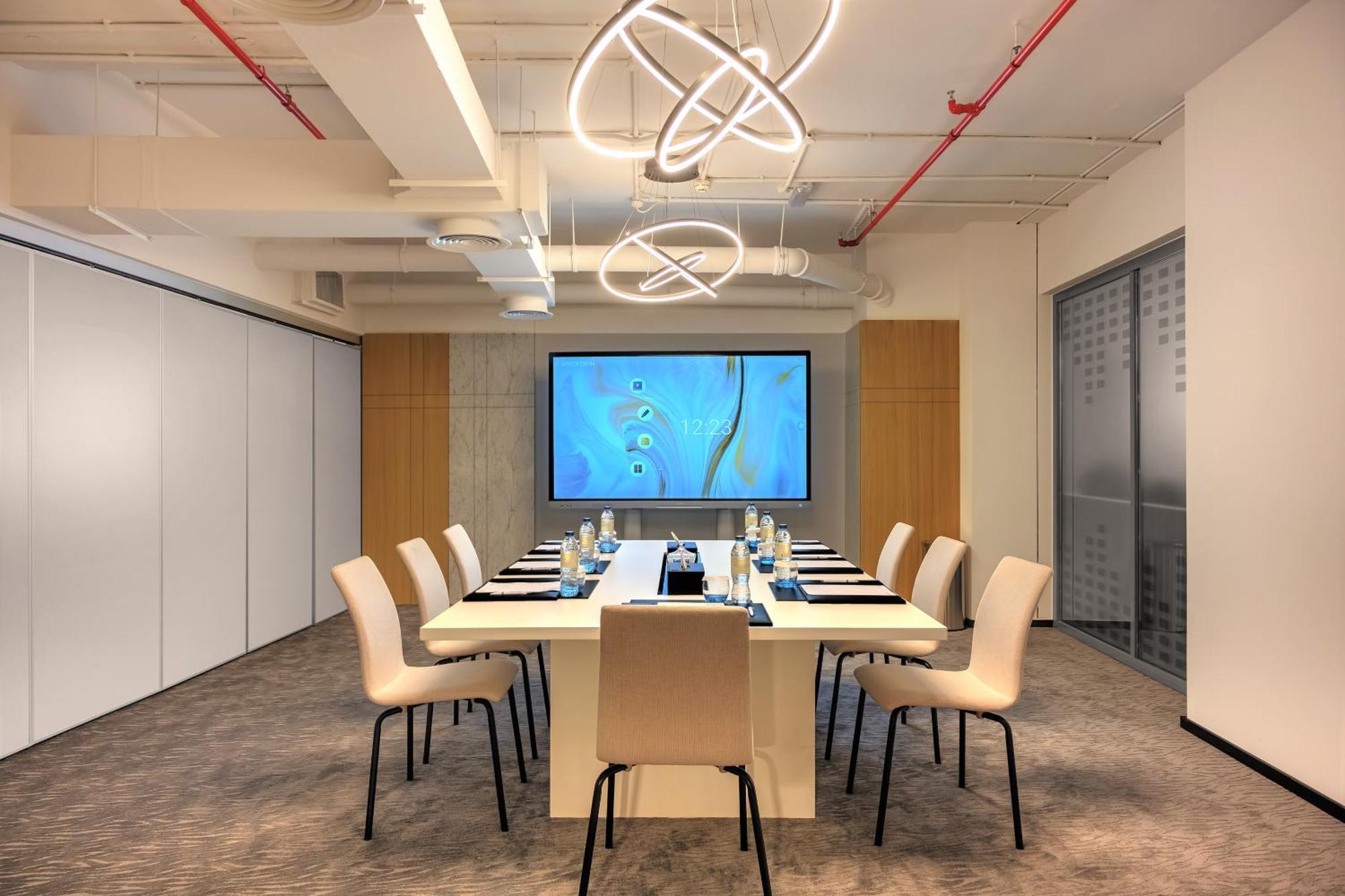 Millennium Atria Business Bay Aparthotel Dubai Luaran gambar The photo shows a modern conference room designed for meetings. It features a long white table surrounded by beige chairs. At the end of the table, there is a large screen displaying a blue abstract design. The room is well-lit with contemporary ligh