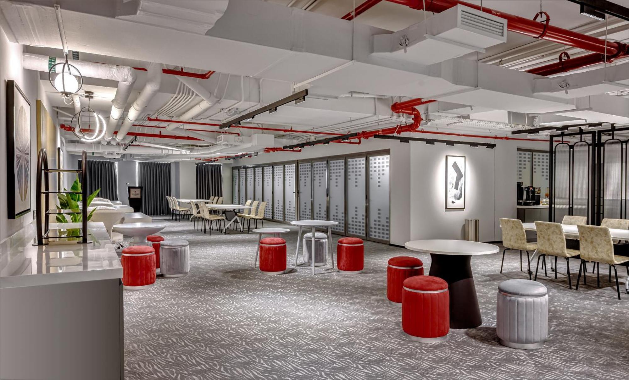 Millennium Atria Business Bay Aparthotel Dubai Luaran gambar The photo shows a modern interior space, likely an office or communal area. The flooring is covered with a textured carpet, and the ceiling features exposed pipes painted red. 

There are several seating options, including small round tables with sto