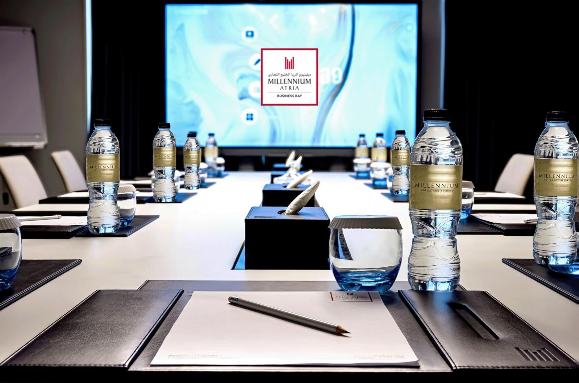 Millennium Atria Business Bay Aparthotel Dubai Luaran gambar The photo shows a modern conference room setup. A long table is laid out with neatly arranged stationery, including notebooks and pens, at each seat. Several bottles of water are placed on the table. In the background, a large screen is displayed, li