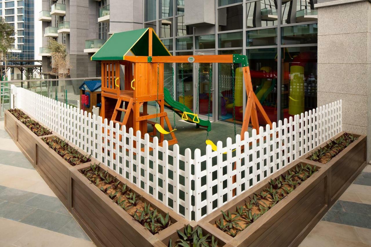 Millennium Atria Business Bay Aparthotel Dubai Luaran gambar The image shows a children's playground area, which is enclosed by a white picket fence. The playground features a wooden play structure with a slide, swings, and climbing elements. Surrounding the play area are planters with greenery, and the backdr