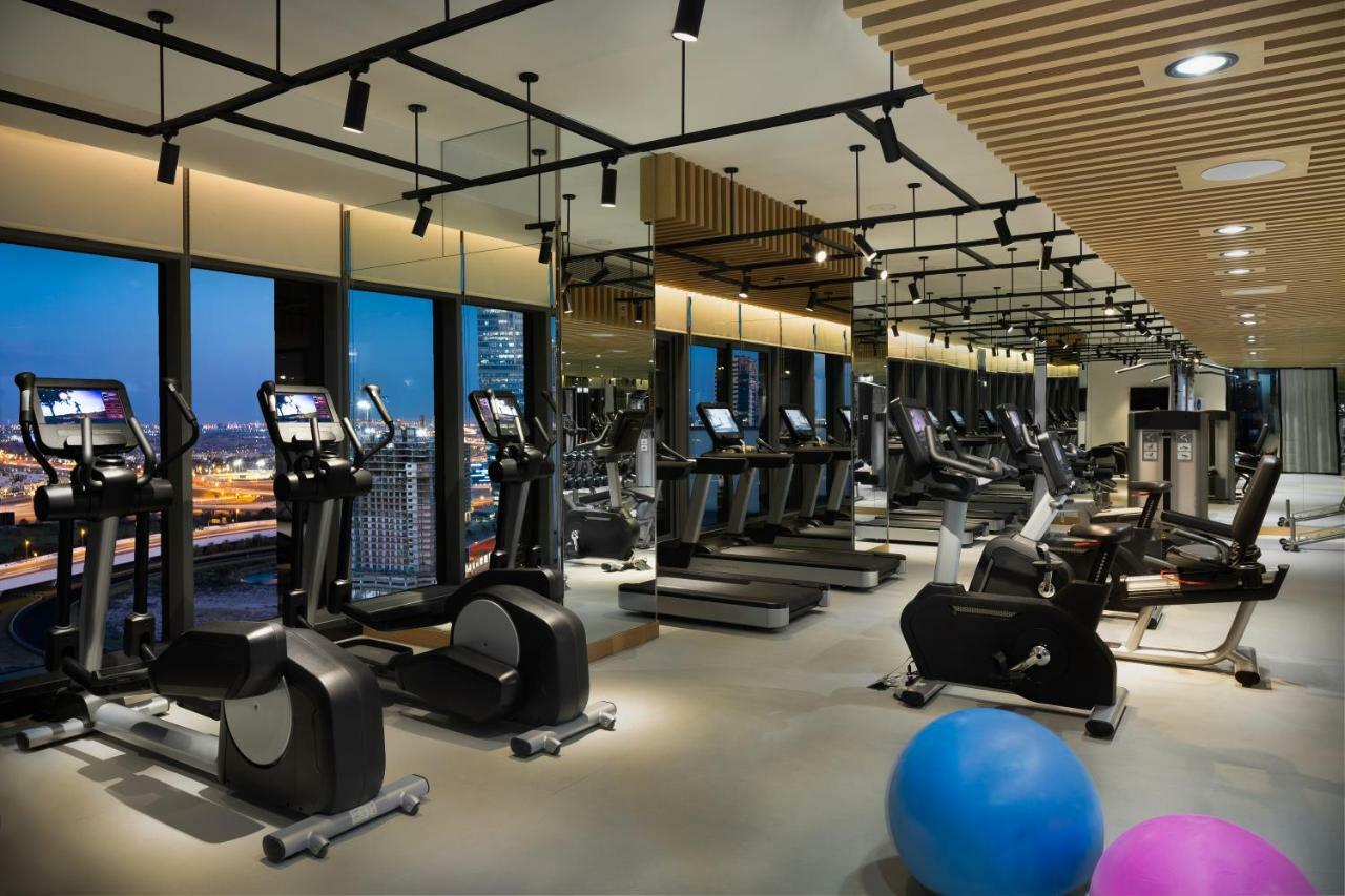 Millennium Atria Business Bay Aparthotel Dubai Luaran gambar The photo shows a modern gym with various types of exercise equipment available. There are several treadmills and elliptical machines arranged in rows, with large windows in the background providing a view of the city. The lighting is bright and cont