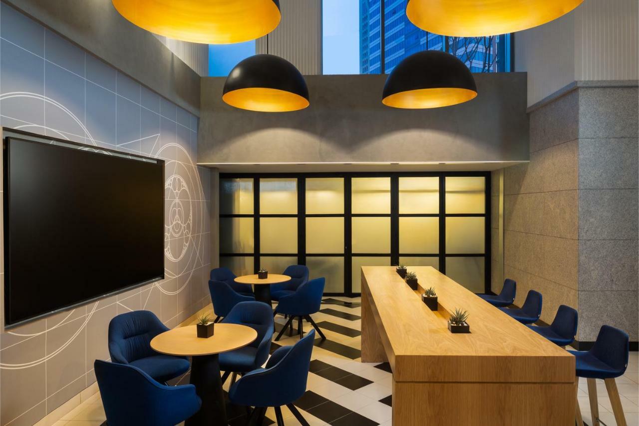 Millennium Atria Business Bay Aparthotel Dubai Luaran gambar The photo shows a modern interior space, likely a lounge or meeting area. It features a long wooden table with some small plants on it, accompanied by several blue, circular chairs arranged around it. There is a large flat-screen television mounted o